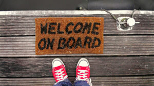 A welcome on board mat on a boat dock with a person in red shoes getting ready to stop onto it.